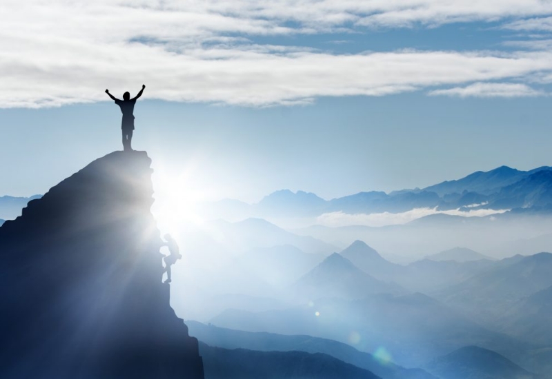 Two people climbing a mountain at sunrise. One stands victorious at the top.