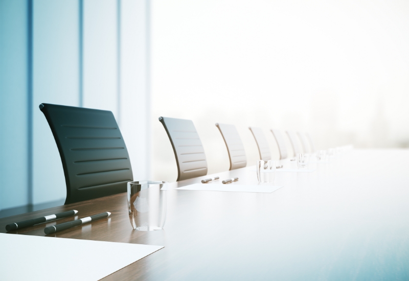 Black boardroom chairs with a piece of paper, a pen and an empty drinking glass in front of each spot at the wooden table.