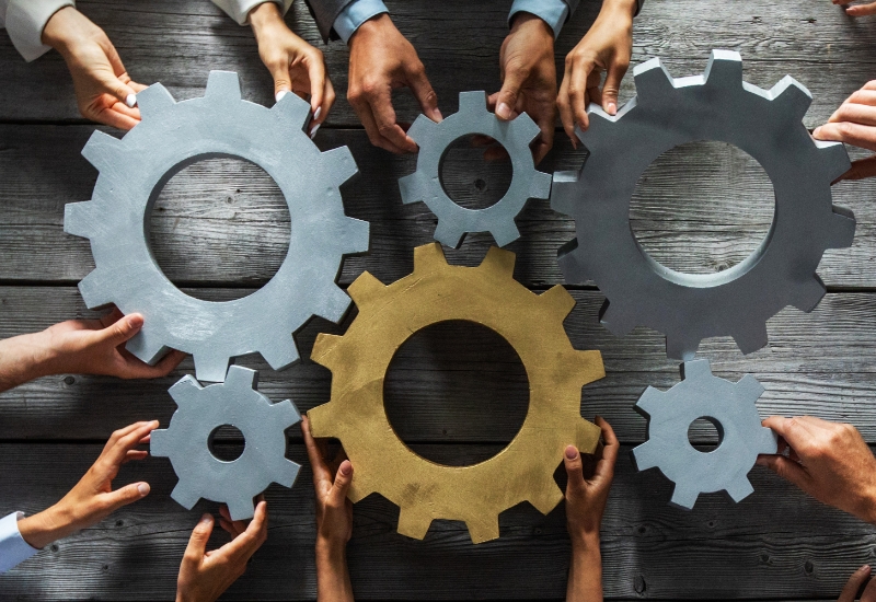 Multiple hands joining silver and gold gears on a wooden tabletop.